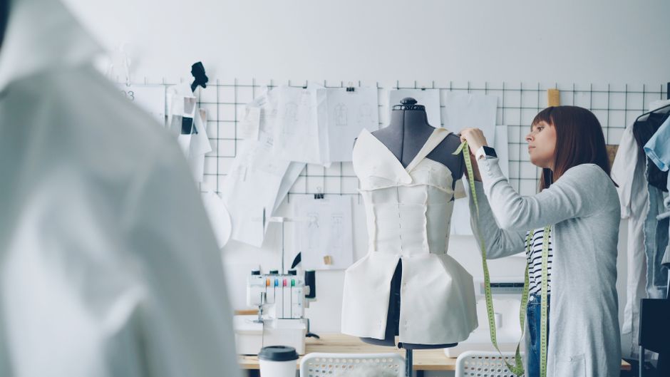 Female designer measuring white dress on mannequin in studio.