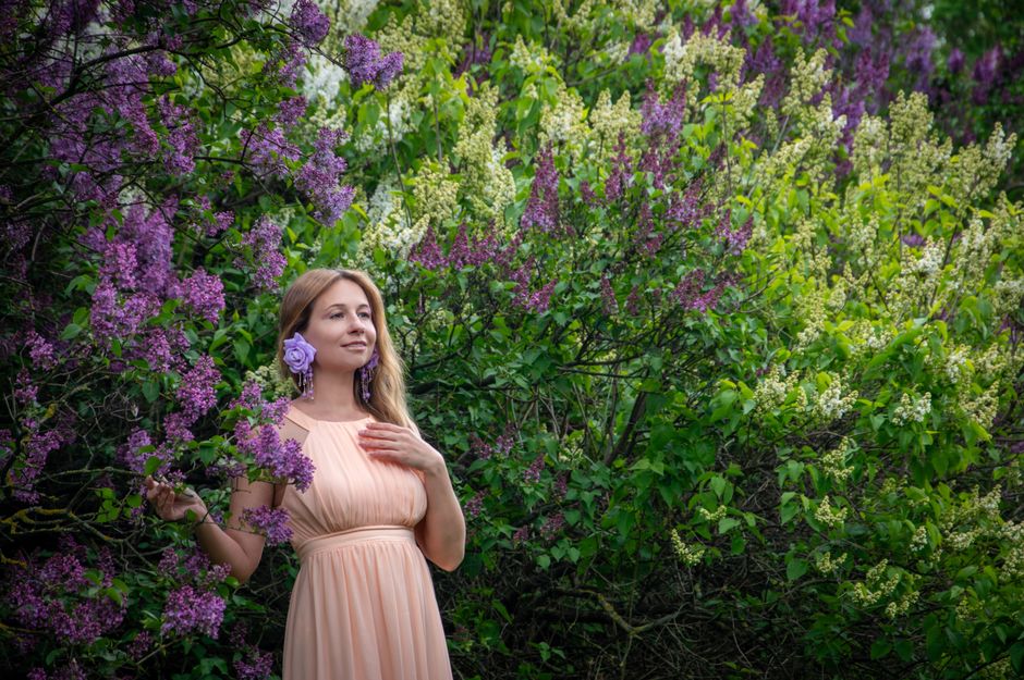 Woman in pastel dress standing gracefully among lush lilac bushes on a spring day.
