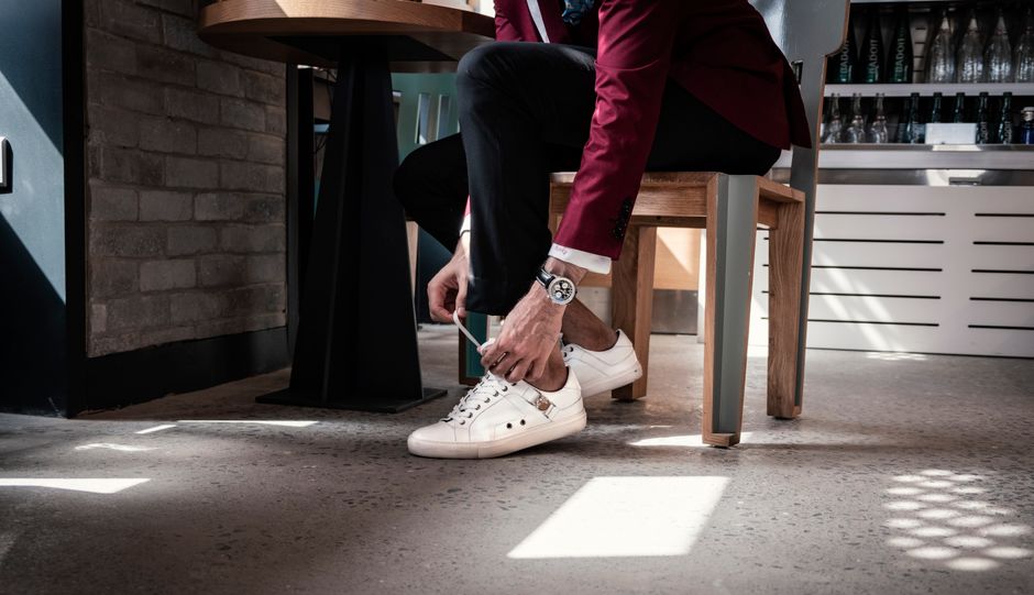 A fashionable man in a burgundy blazer tying shoelaces in a modern indoor setting.